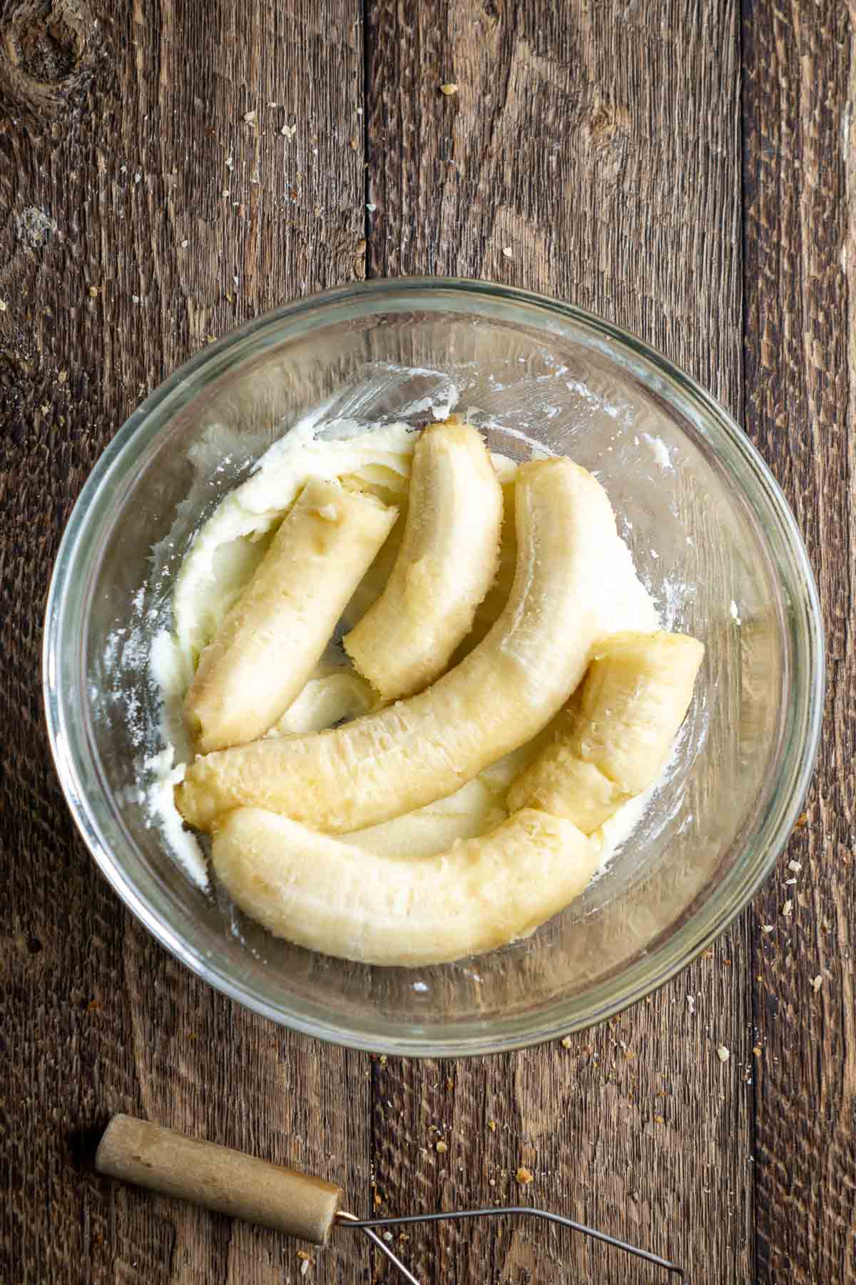 Peeled bananas in a glass mixing bowl with creamed butter and sugar, on a wooden surface. A wire whisk is nearby.