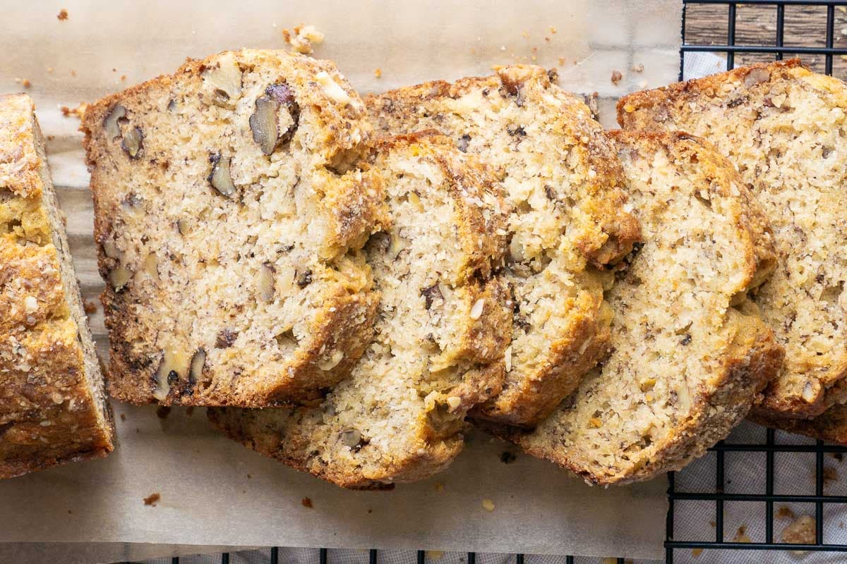 A partially sliced loaf of banana bread with visible nuts rests on parchment paper atop a black wire cooling rack, highlighting the moist texture and classic appeal of homemade banana bread.