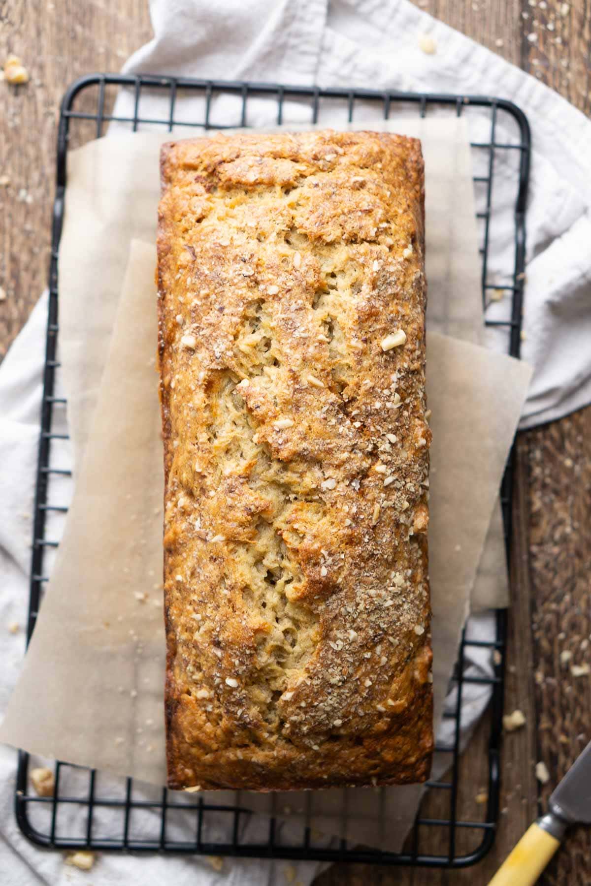 A loaf of homemade banana bread with a golden-brown crust sits on parchment paper atop a cooling rack.
