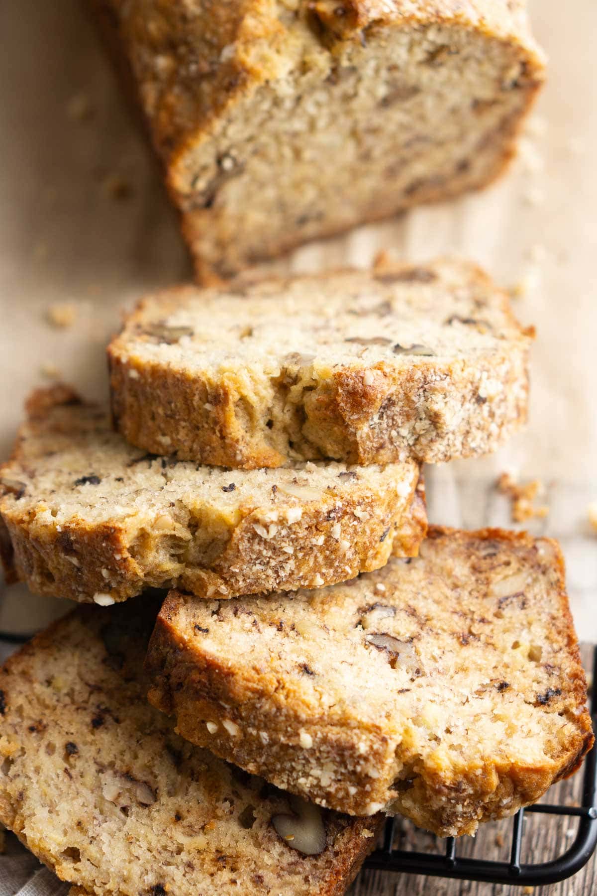 Close-up of several slices of golden brown banana walnut bread pieces, stacked on a cooling rack in front of a fresh banana nut bread loaf.