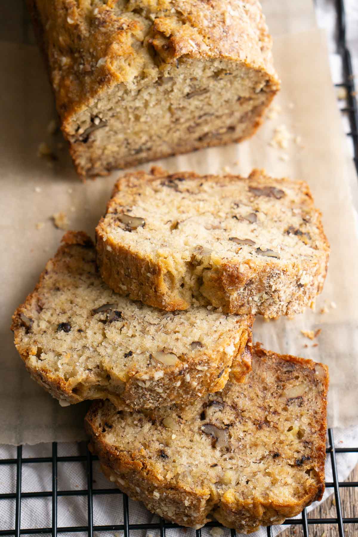 Close-up of several slices of golden brown banana walnut bread pieces, stacked on a cooling rack in front of a fresh banana nut bread loaf.