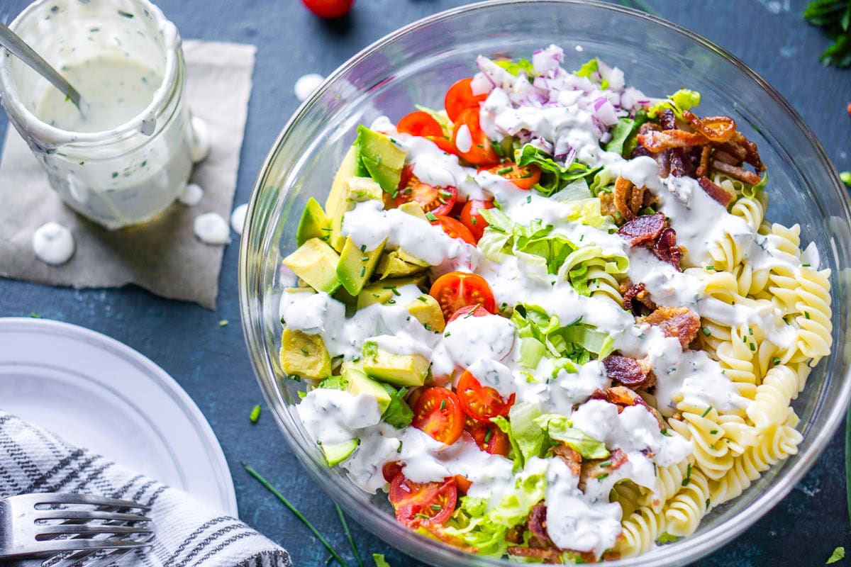 A glass bowl filled with blt pasta salad topped with creamy white dressing next to jar of dressing and dinnerware.
