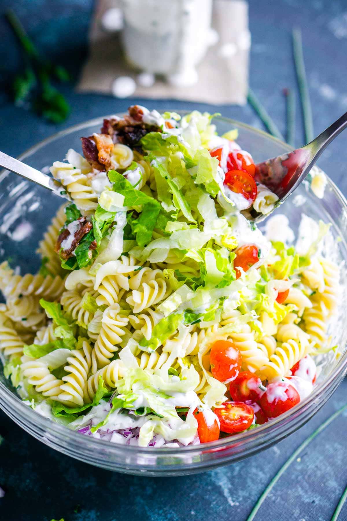 A clear glass bowl filled with blt pasta salad with two serving utensils in the bowl picking up the salad.