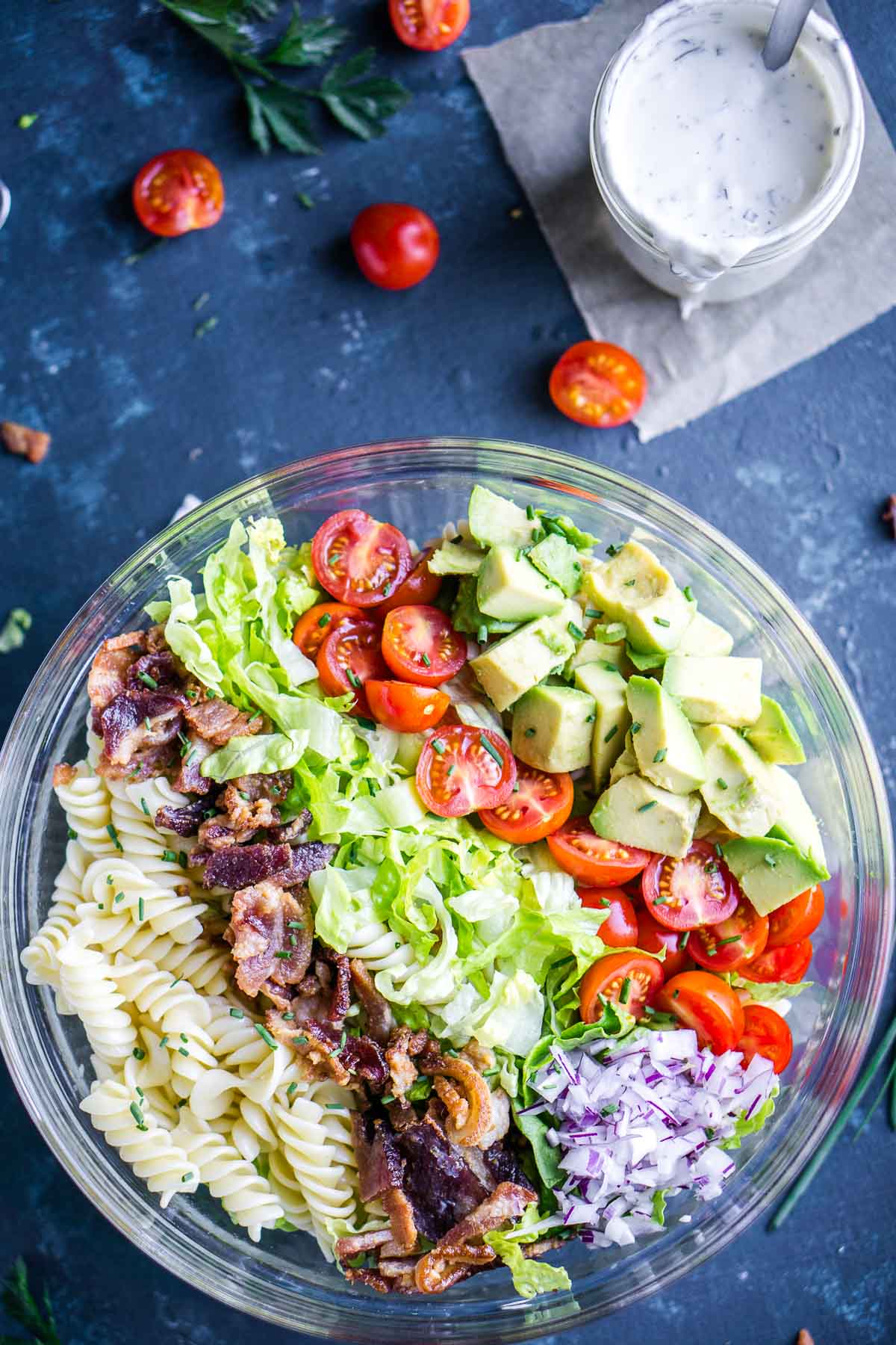 A glass bowl filled with blt pasta salad next to jar of creamy white dressing and a few halved cherry tomatoes.