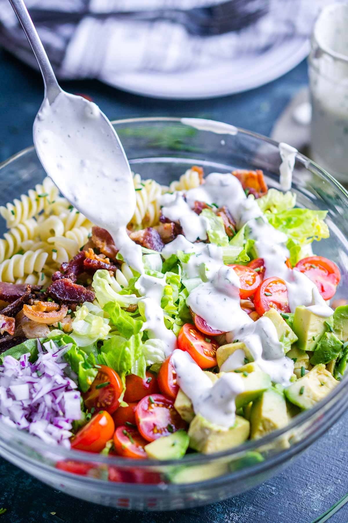 A glass bowl filled with blt pasta salad being drizzled with creamy white dressing from a spoon.