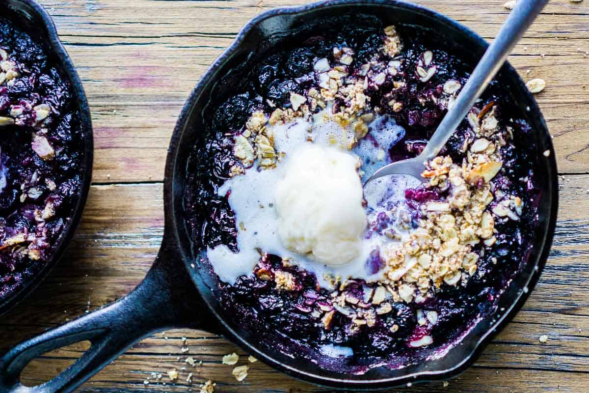 Ice cream is melted over warm blueberry crisp in iron skillet with serving spoon
