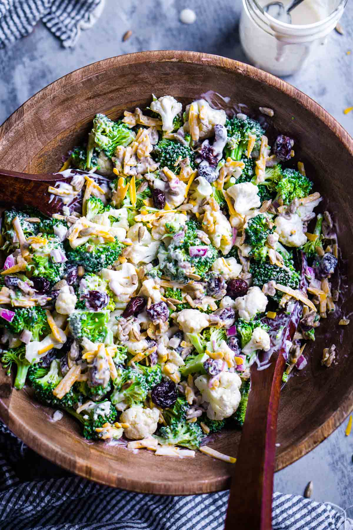 broccoli cauliflower salad in wood bowl with salad serving utensils next to dressing jar