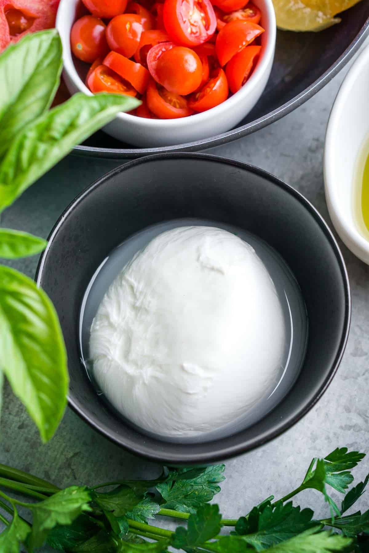 burrata cheese in black bowl next to tomatoes and fresh herbs