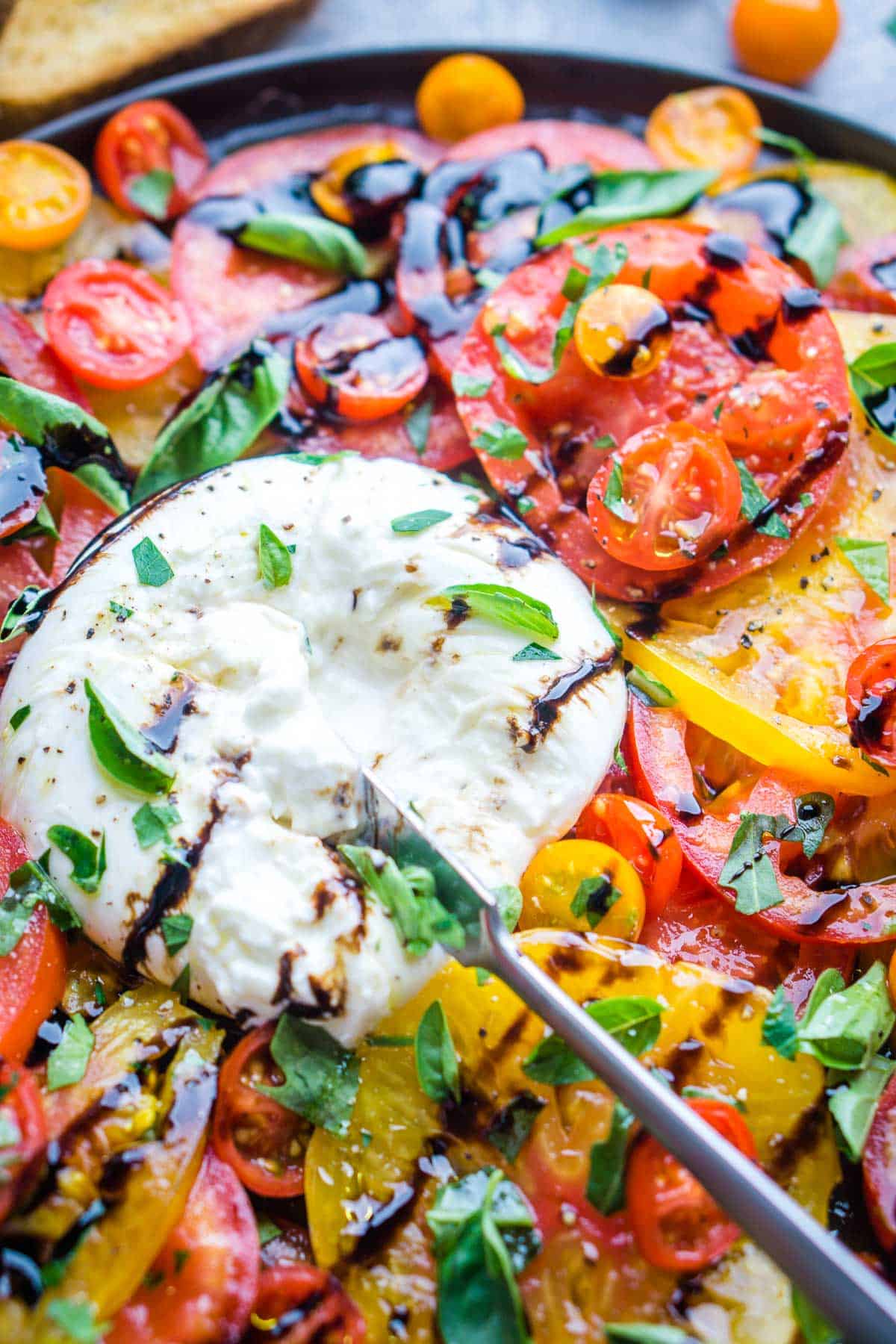 knife cuts into burrata cheese surrounded by tomatoes on gray plate