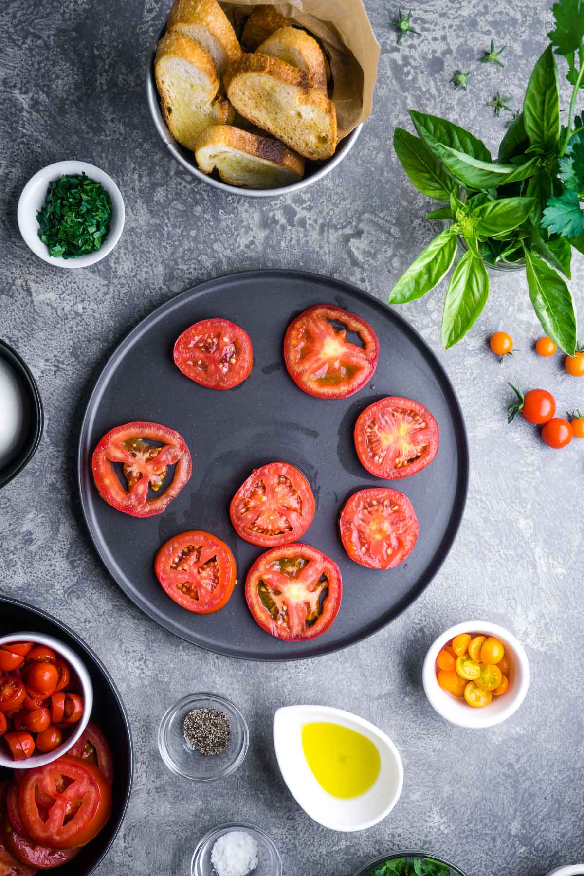 Sliced tomatoes on a black plate surrounded by bread, herbs, whole cherry tomatoes, olive oil, salt, pepper, and chopped parsley on a gray surface.