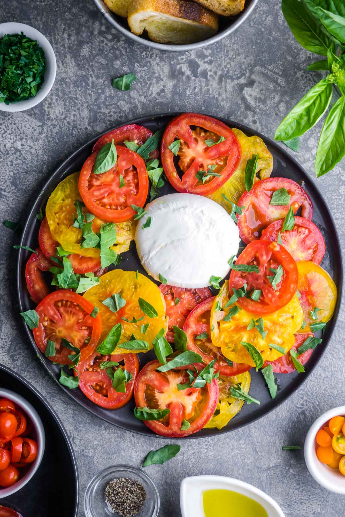 A plate with sliced red and yellow tomatoes, fresh basil leaves, and a ball of mozzarella cheese in the center, reminiscent of a classic burrata salad, surrounded by bowls of herbs, cherry tomatoes, and olive oil.