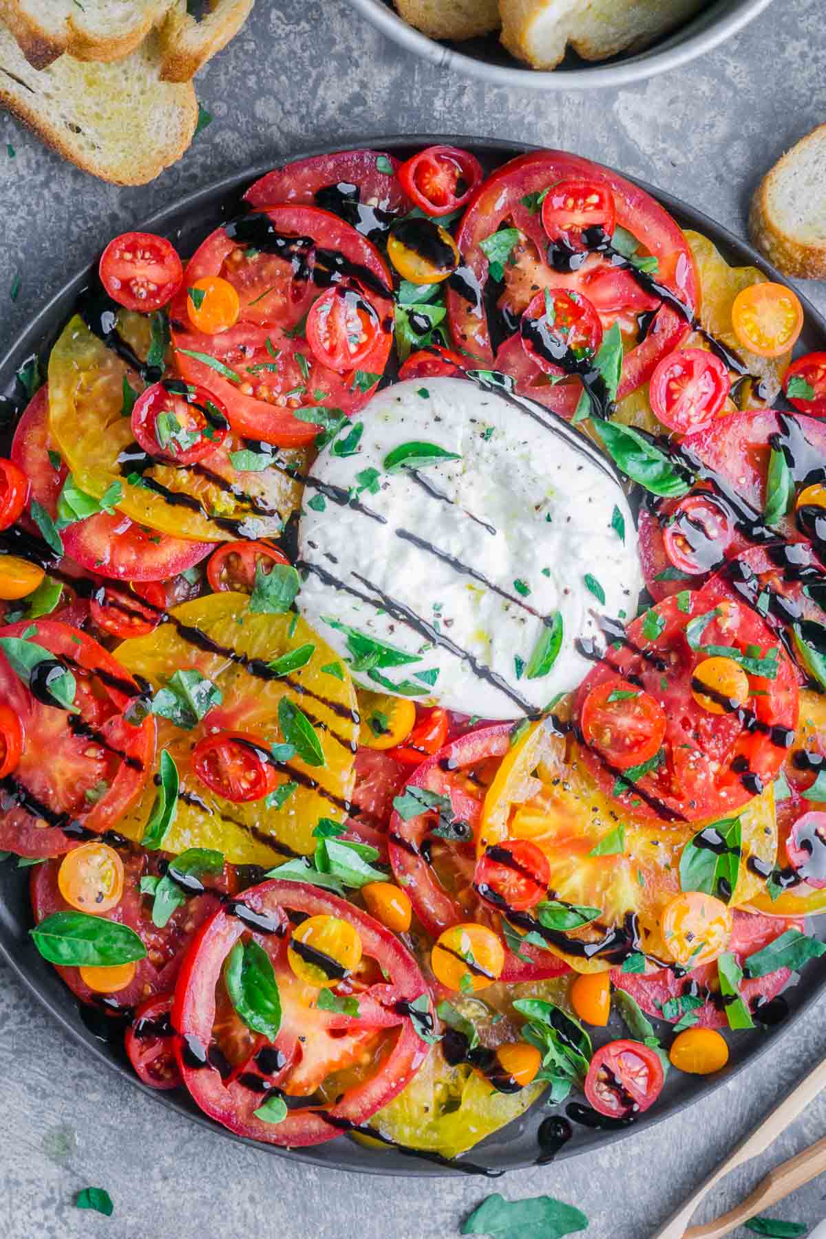 Tomato burrata salad layers juicy heirloom and cherry tomatoes with creamy burrata, fresh basil, olive oil, and balsamic glaze. This easy summer appetizer or side dish comes together in minutes and is always a crowd favorite. Colorful sliced cherry tomatoes and heirloom tomatoes surround a ball of fresh burrata, topped with basil and balsamic glaze, on a round black plate for an elegant burrata salad. Bread slices are visible in the background.