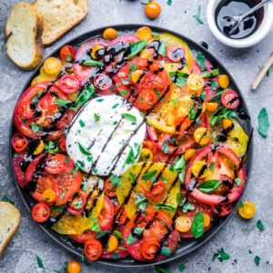 Colorful sliced heirloom tomatoes surround a ball of fresh burrata, topped with basil and balsamic glaze, on a round black plate for an elegant burrata salad. Bread slices are visible in the background.
