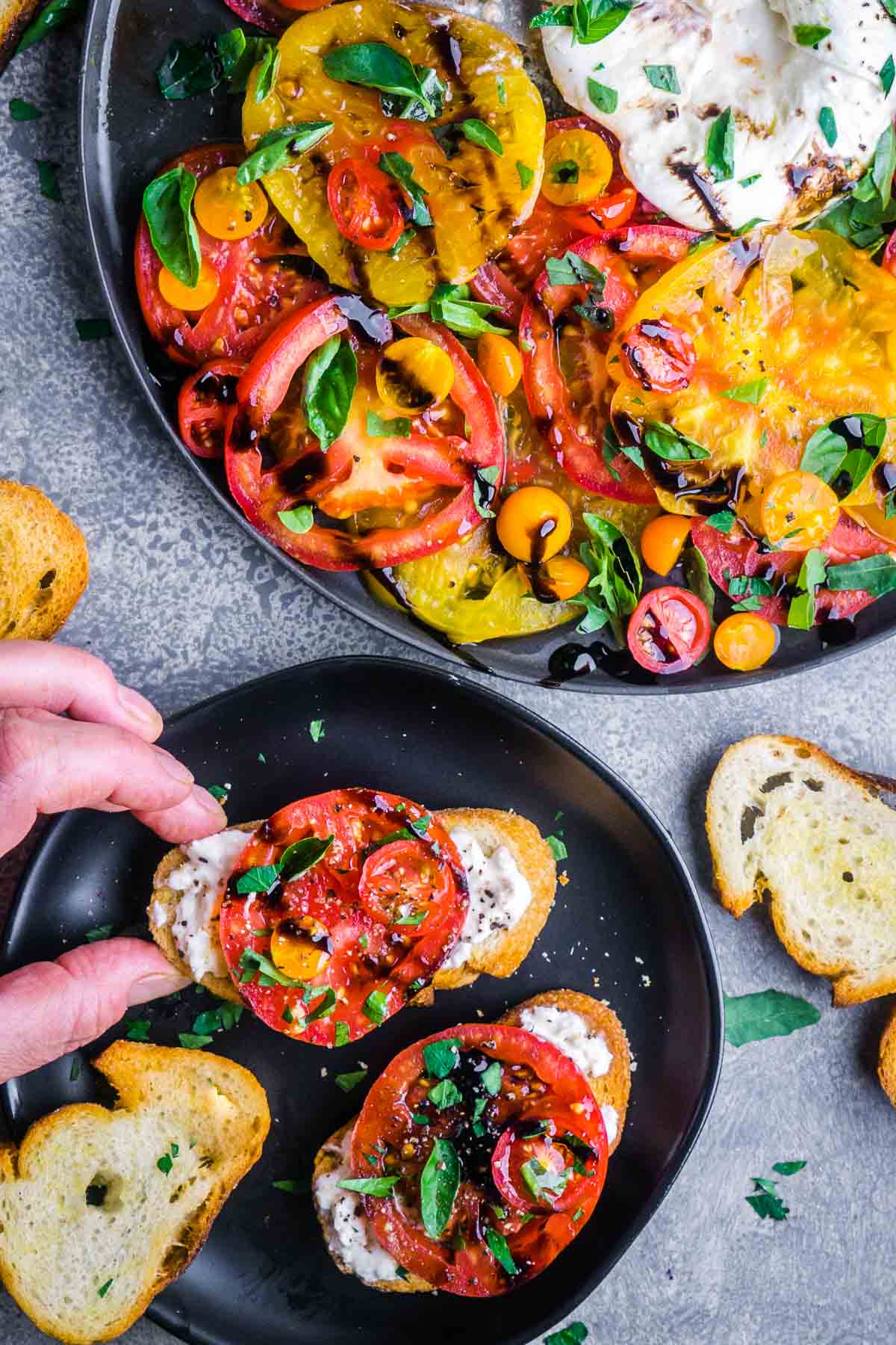 Person's hand picking up tomato burrata salad on crostini from a platter with creamy burrata, fresh basil, and balsamic glaze.