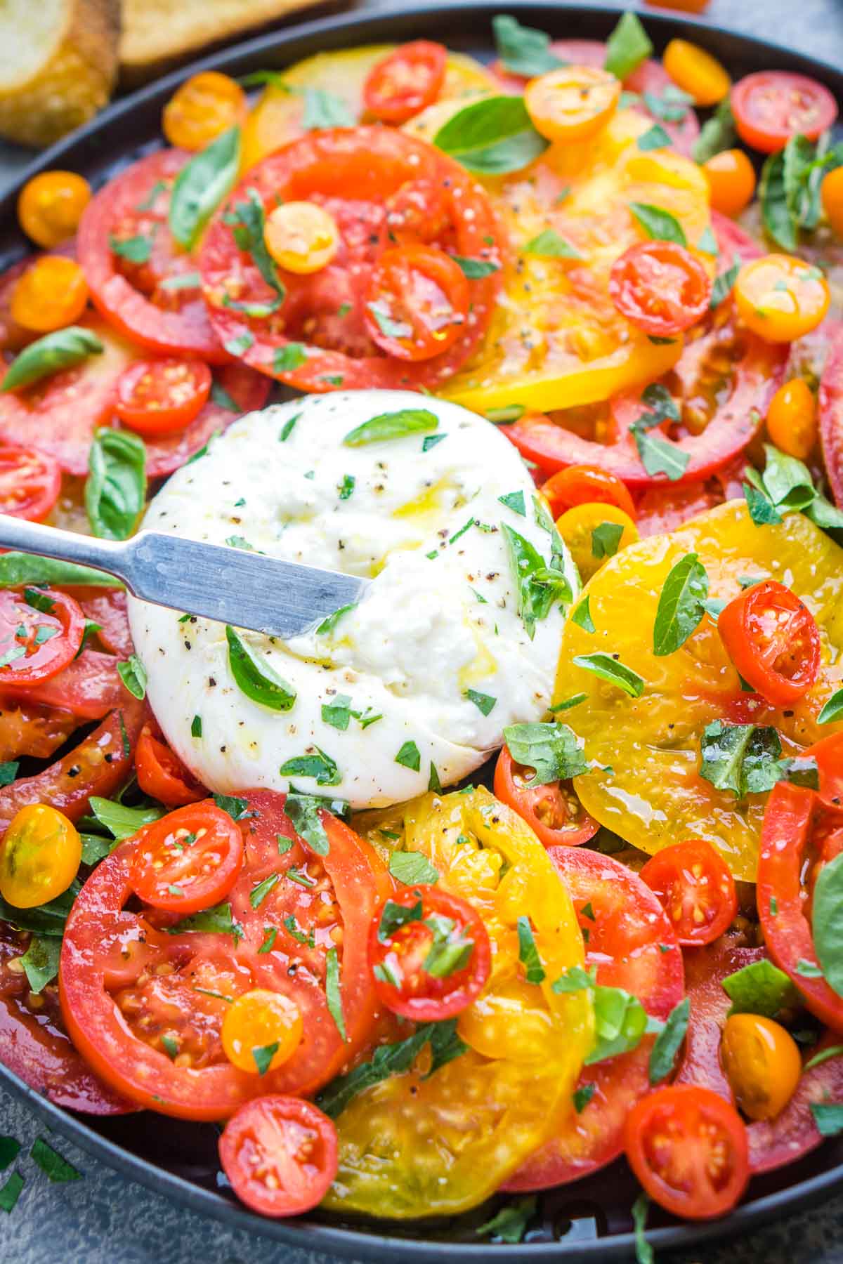 A vibrant burrata salad featuring a plate of sliced red and yellow tomatoes, juicy cherry tomatoes, fresh basil, and a creamy ball of burrata cheese being opened with a knife.