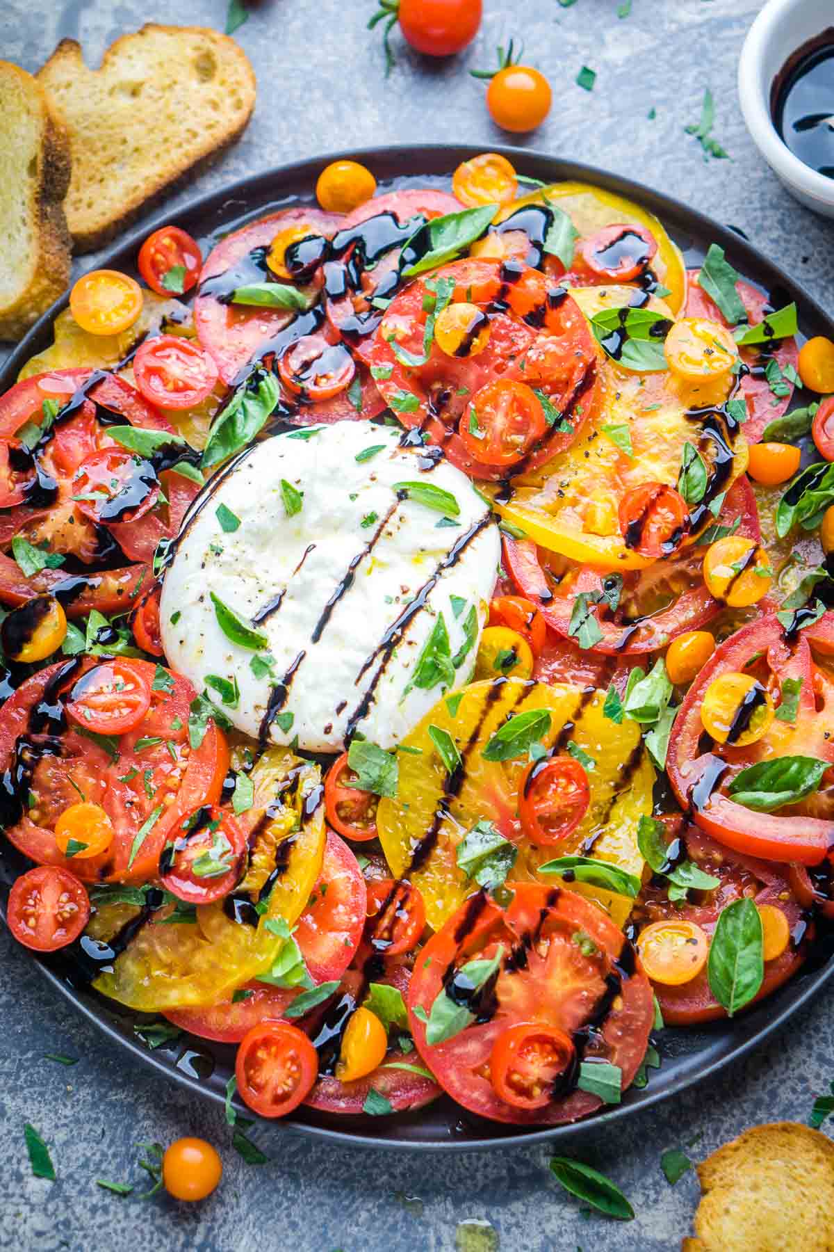 Colorful sliced heirloom tomatoes and cherry tomatoes surround a ball of fresh burrata, topped with basil and balsamic glaze, on a round black plate for an elegant burrata salad. Bread slices are visible in the background.