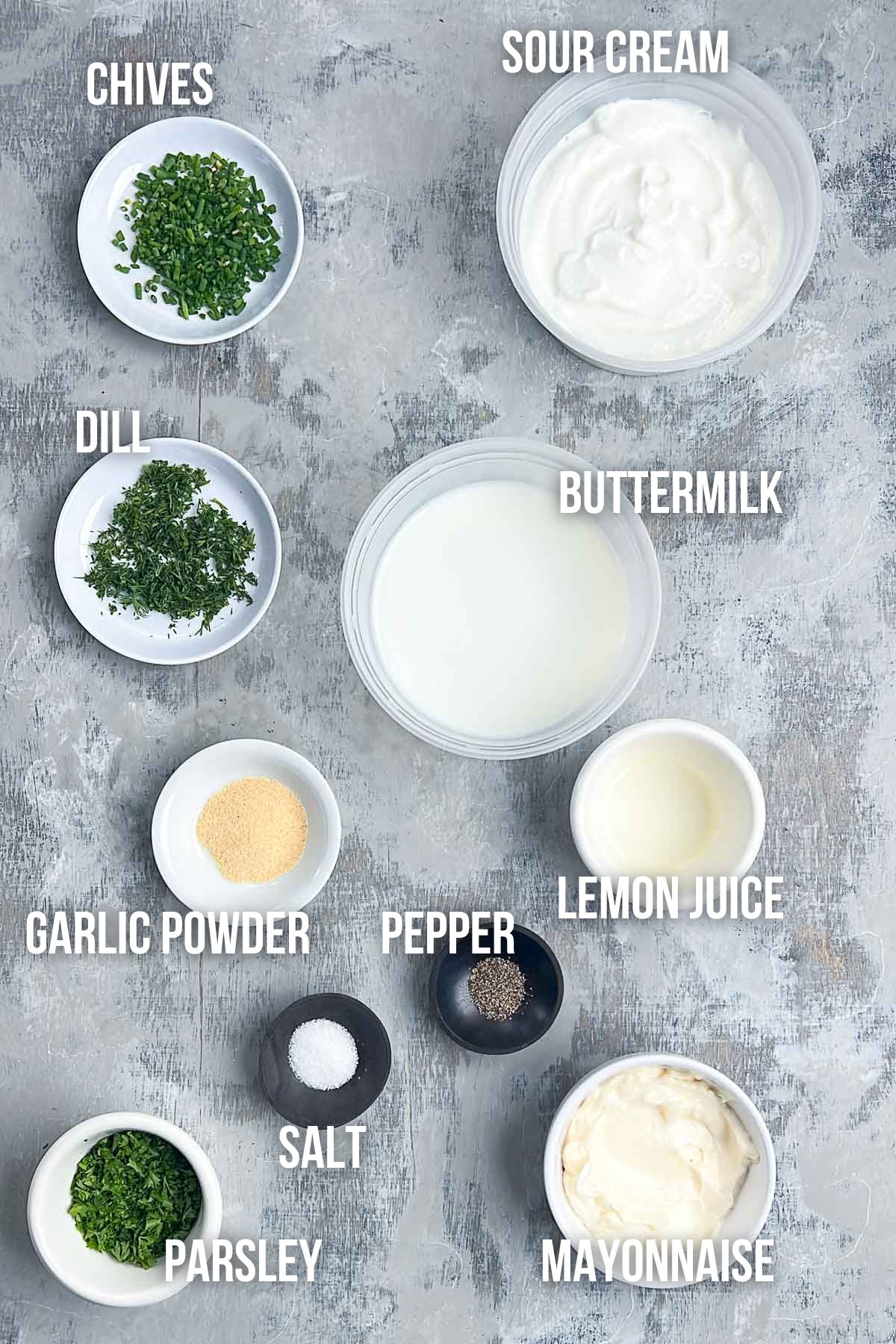 Various ingredients for buttermilk ranch dressing, including chopped chives, dill, parsley, garlic powder, salt, pepper, lemon juice, buttermilk, sour cream, and mayonnaise, are arranged in bowls.