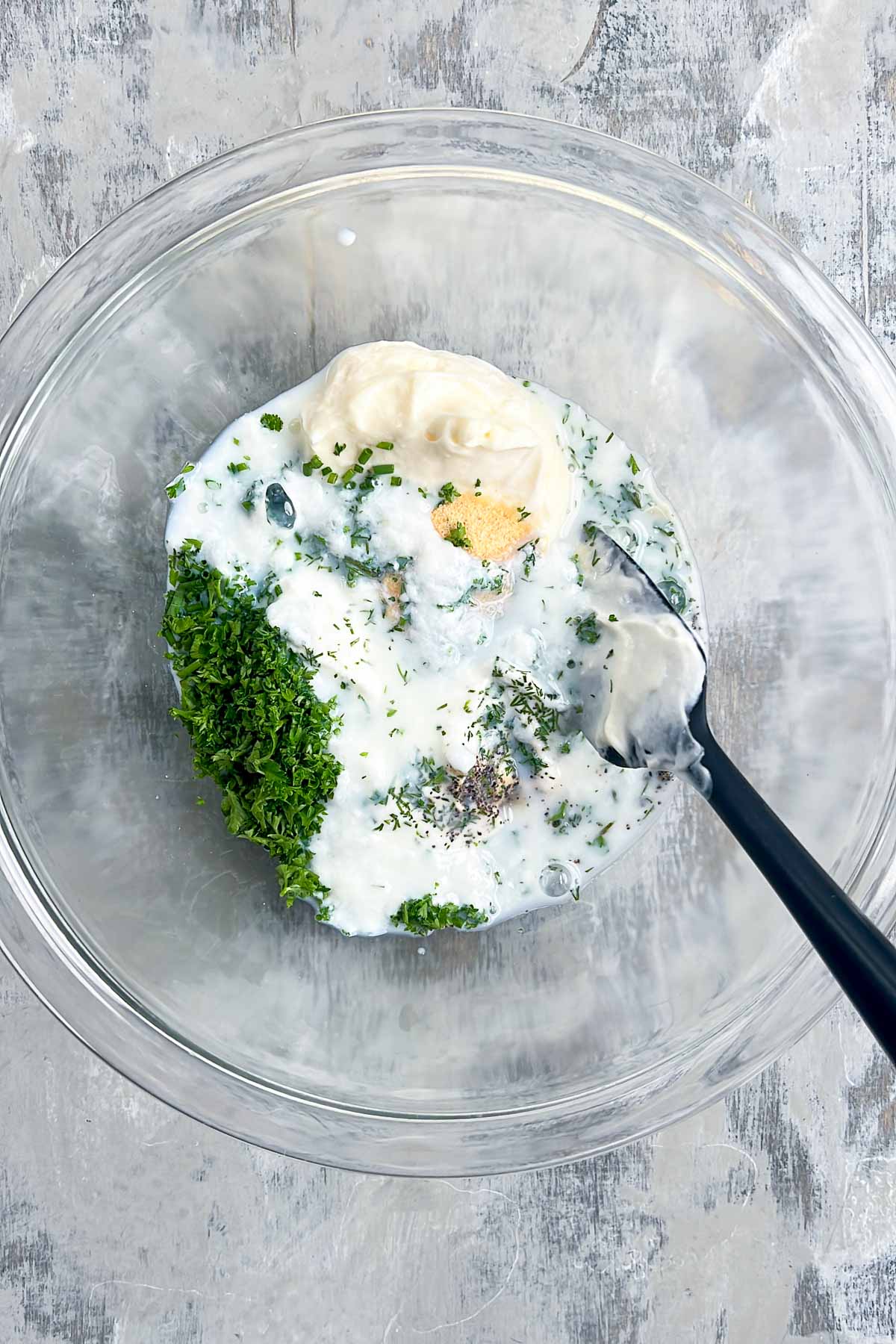 A glass bowl containing the ingredients for buttermilk ranch dressing—yogurt, mayonnaise, chopped herbs, dried seasonings, and a spoon—ready to be mixed.