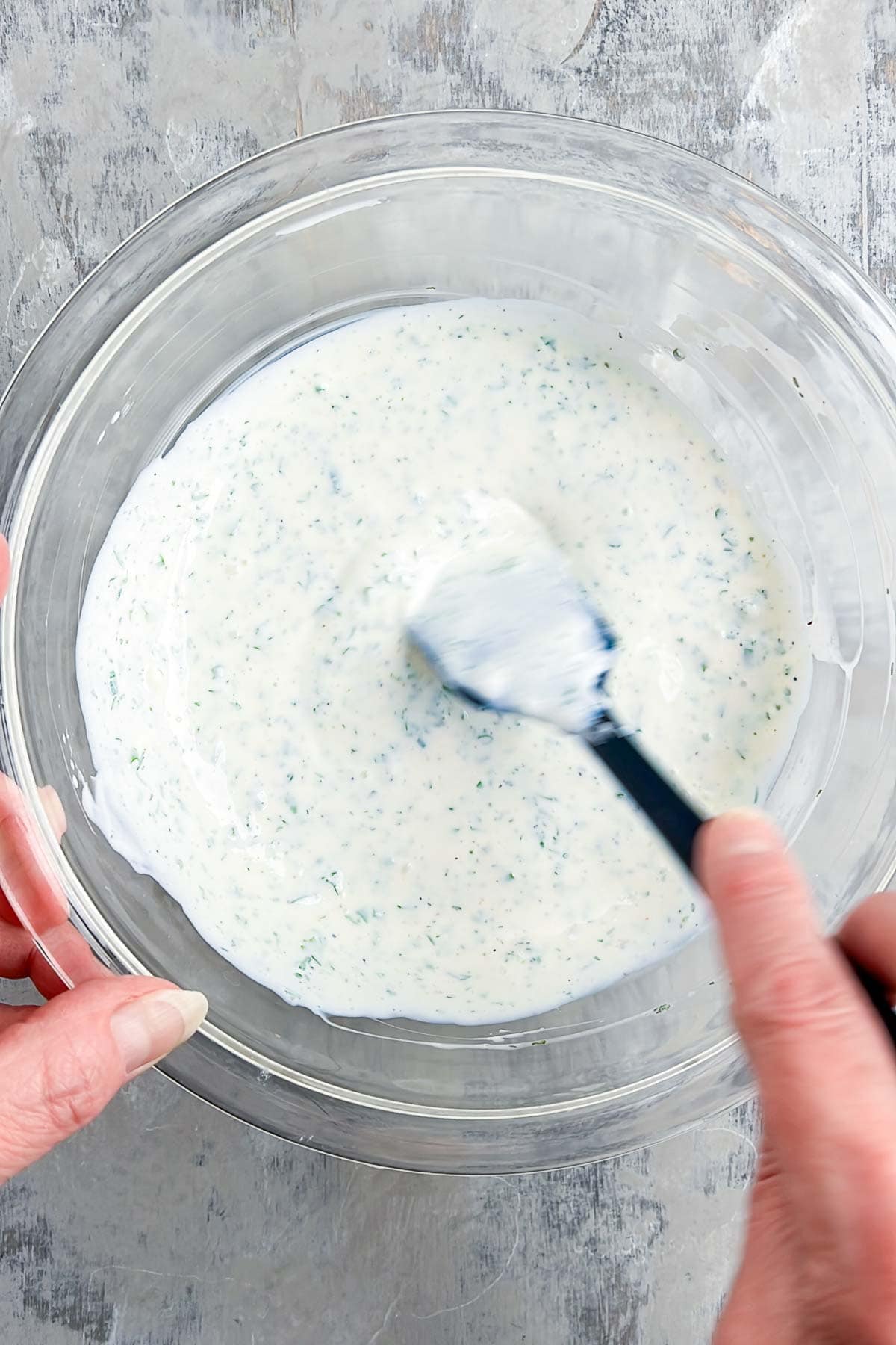 A person stirs a creamy buttermilk ranch dressing with green herbs in a clear glass bowl using a spatula.