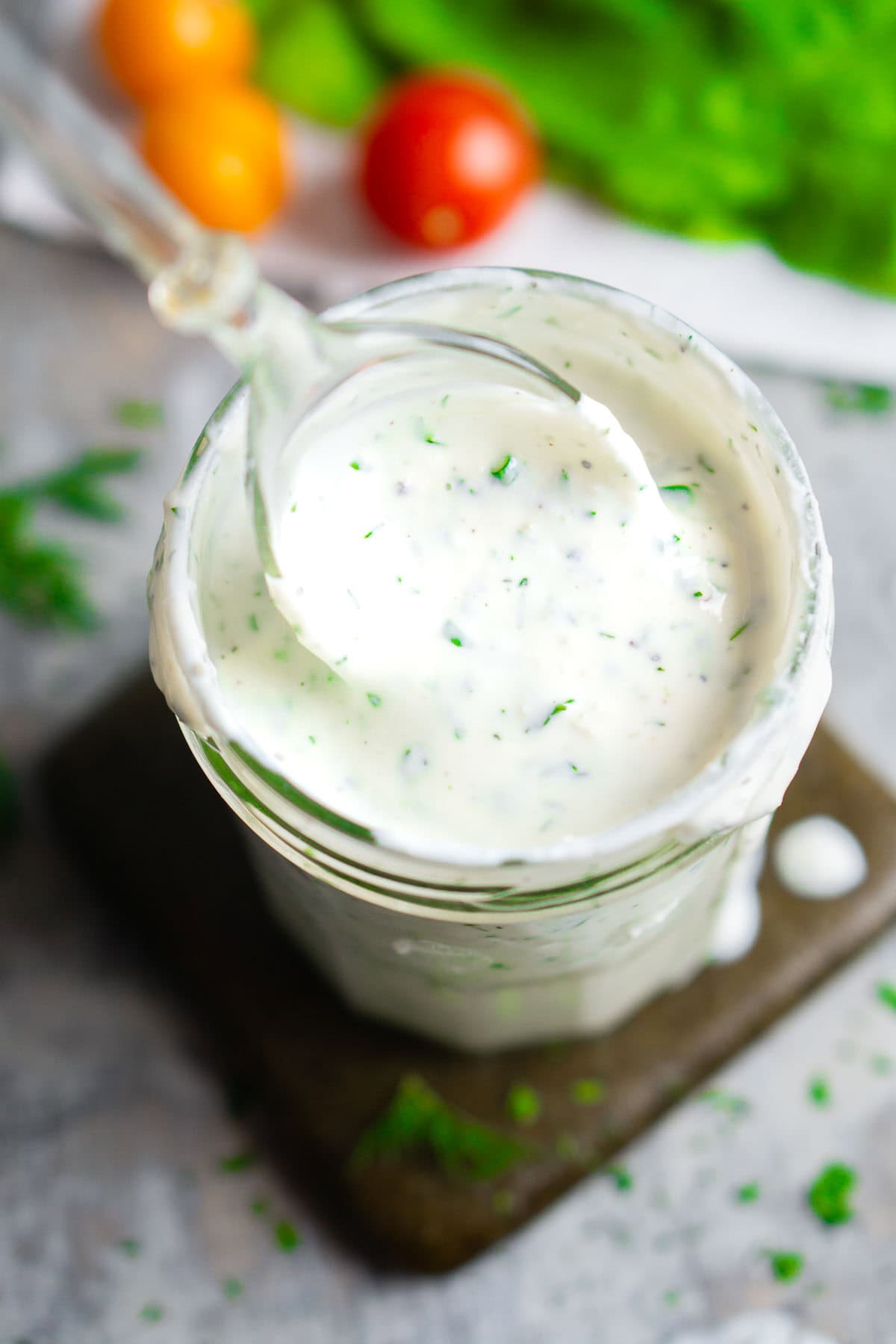 A glass jar filled with creamy buttermilk ranch dressing, topped with herbs, with a spoon resting inside. Fresh tomatoes and leafy greens are blurred in the background.