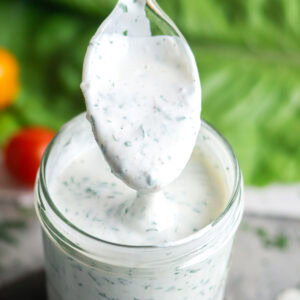 A spoonful of creamy, herb-speckled buttermilk ranch dressing is held above a glass jar filled with the same dressing, with fresh greens and tomatoes in the background.