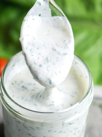 A spoonful of creamy, herb-speckled buttermilk ranch dressing is held above a glass jar filled with the same dressing, with fresh greens and tomatoes in the background.