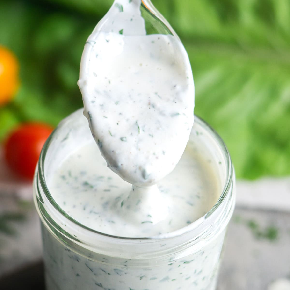 A spoonful of creamy, herb-speckled buttermilk ranch dressing is held above a glass jar filled with the same dressing, with fresh greens and tomatoes in the background.