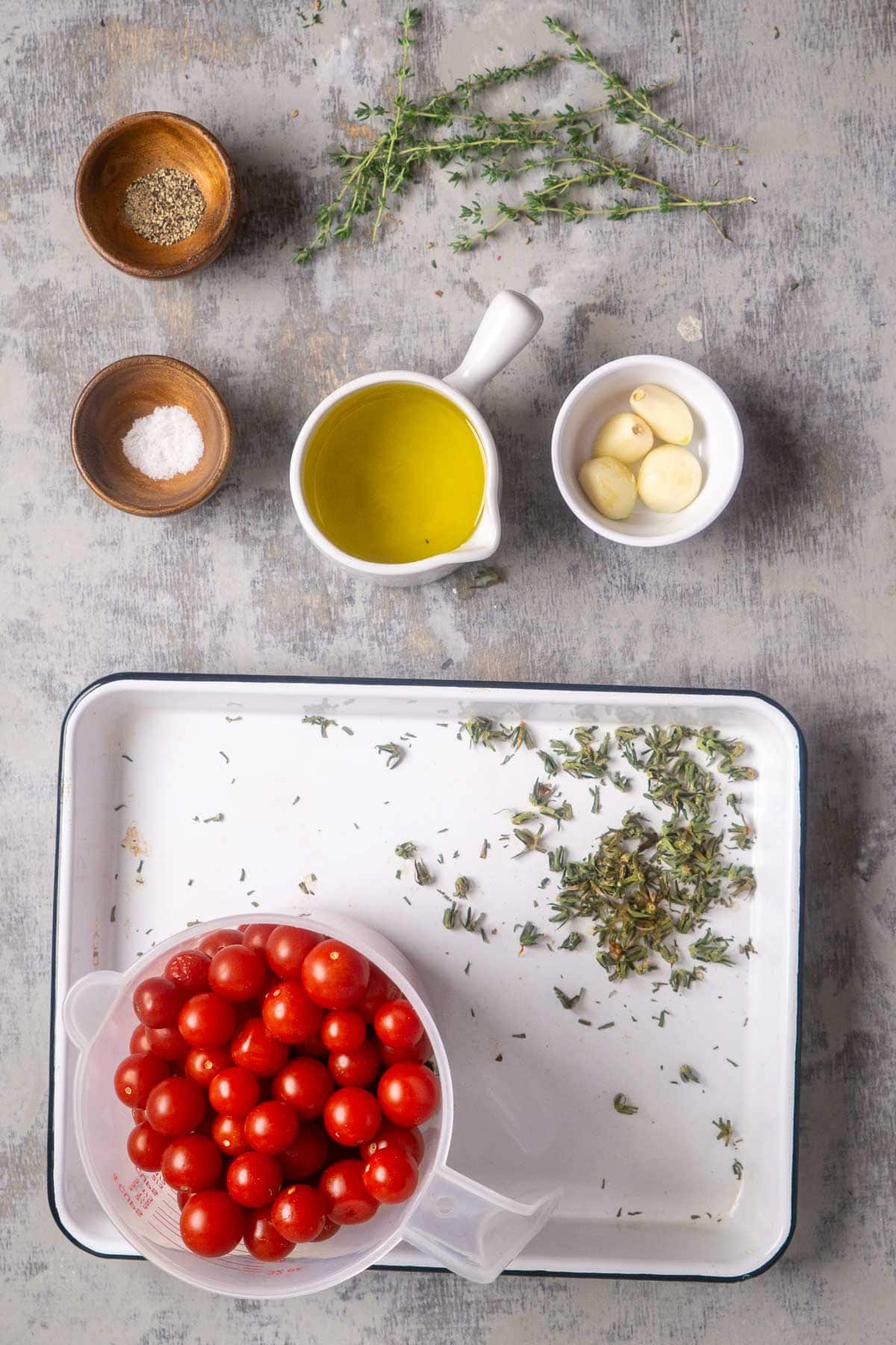 A tray with cherry tomatoes, fresh herbs, olive oil, garlic cloves, salt, and pepper arranged on a gray surface.