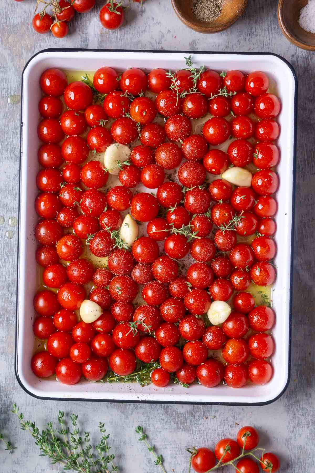 A tray with cherry tomatoes, fresh herbs, olive oil, garlic cloves, salt, and pepper arranged on a gray surface.