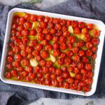 A white baking dish filled with roasted cherry tomatoes, garlic cloves, olive oil, and fresh herbs on a dark cloth.