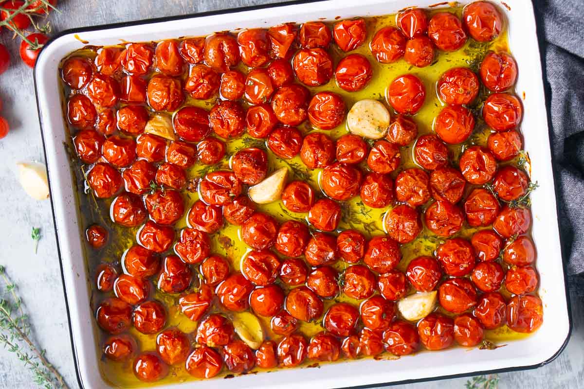 A white baking dish filled with roasted cherry tomatoes, garlic cloves, olive oil, and fresh herbs on a dark cloth.
