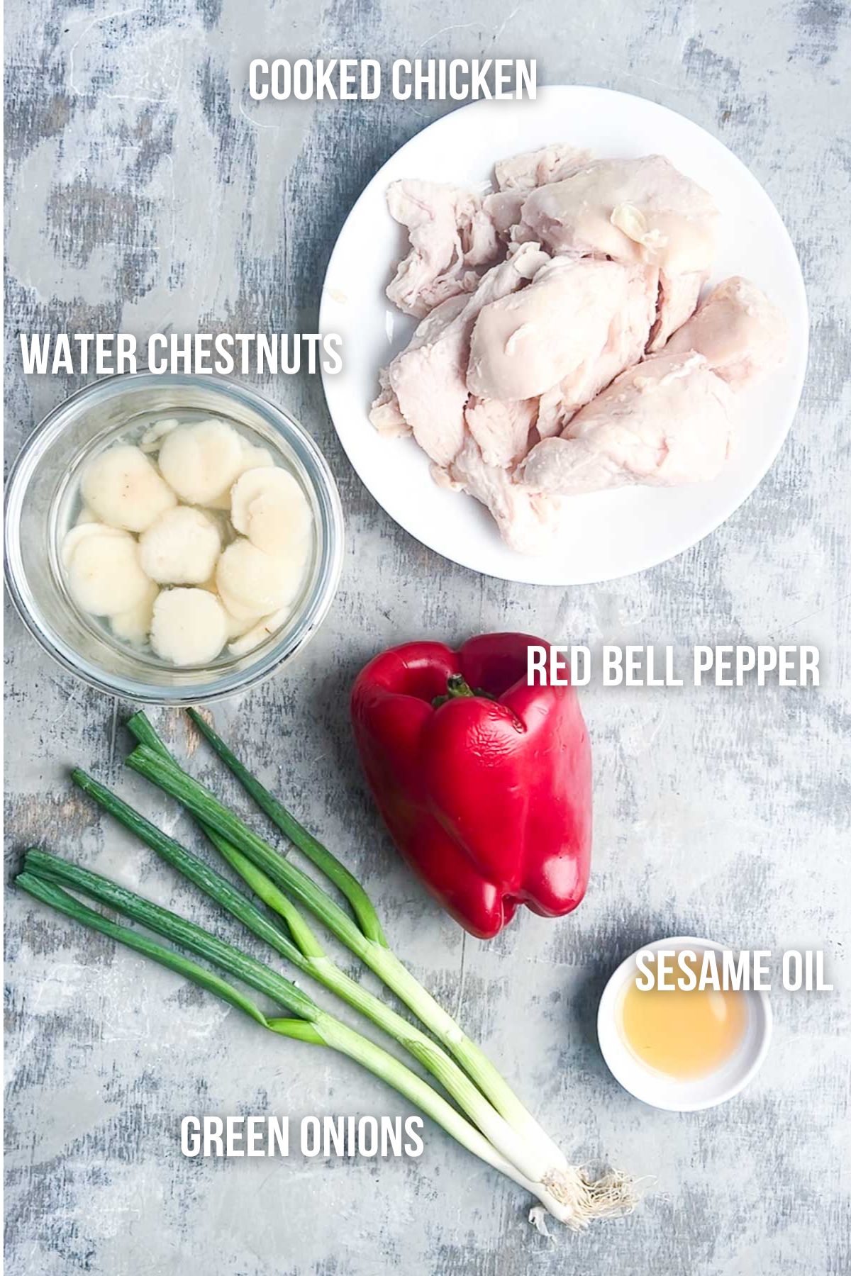 A plate of cooked chicken, a bowl of water chestnuts, a red bell pepper, green onions, and a small dish of sesame oil arranged on a gray surface&mdash;ingredients needed to make chicken lettuce wraps.