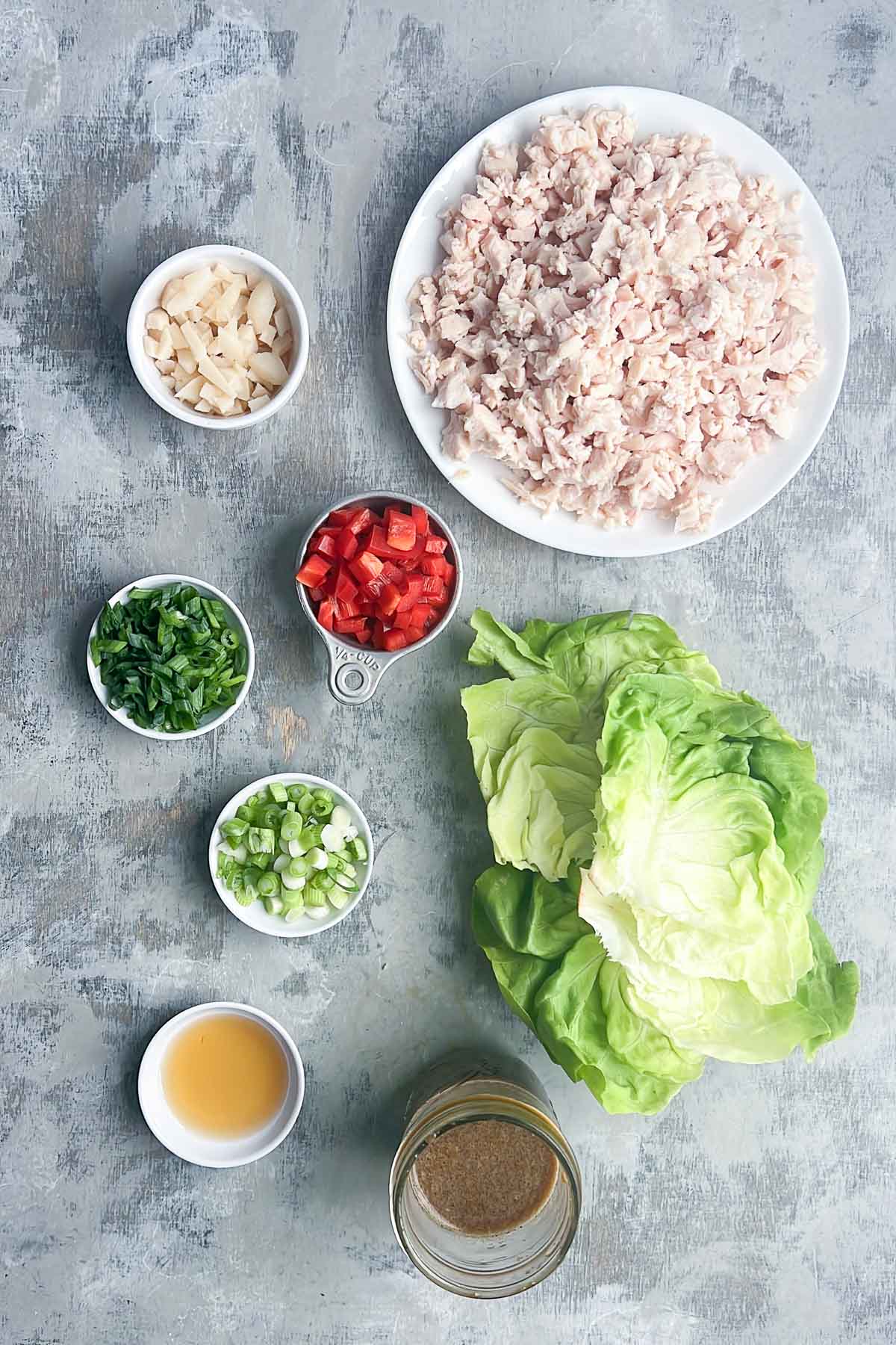 Overhead view of ingredients for chicken lettuce wraps: chopped cooked chicken, diced red bell pepper, sliced green onions, chopped water chestnuts, fresh lettuce leaves, a small cup of sauce, and a jar of brown dressing on a gray surface.