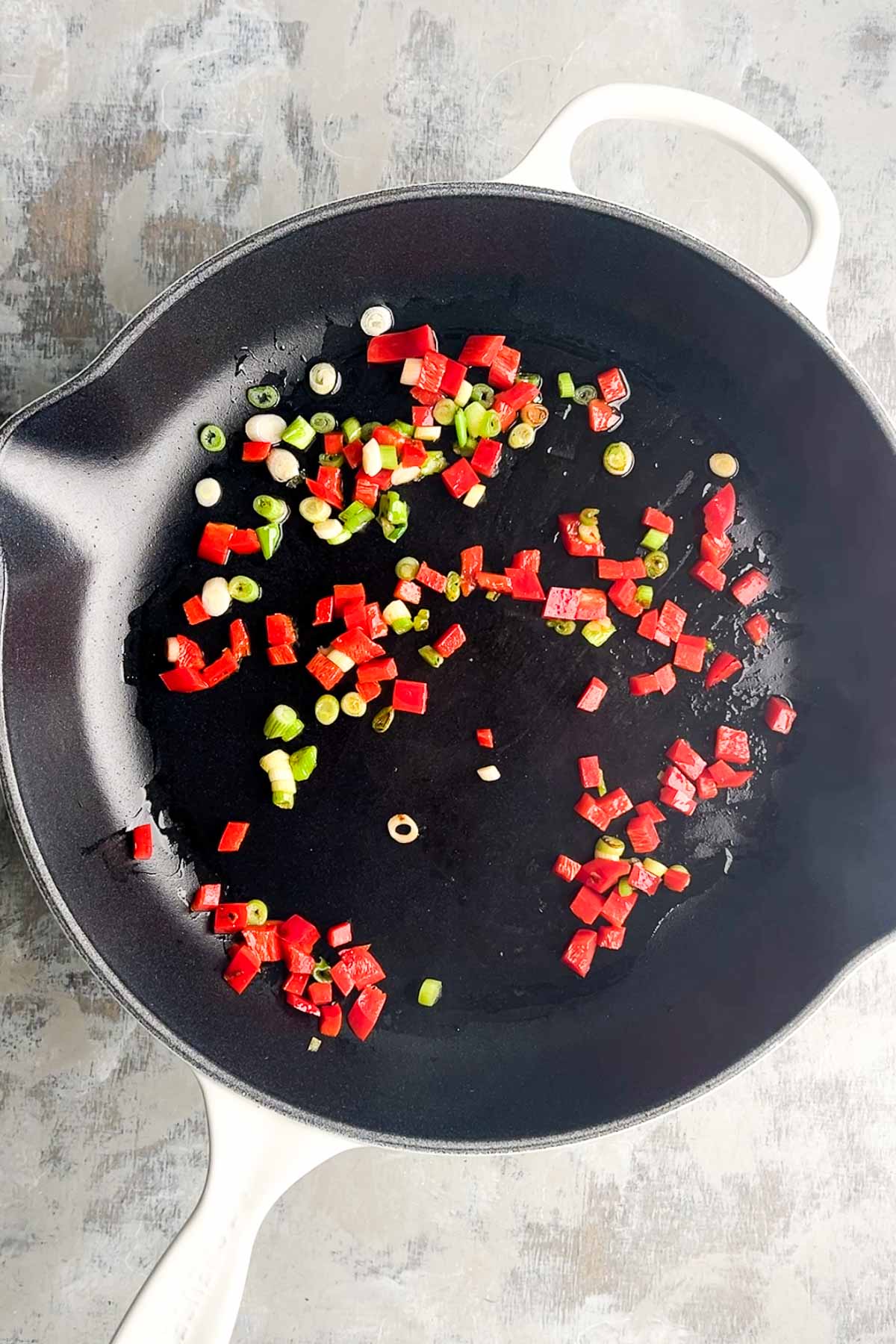Diced red bell pepper and chopped green onions saut&eacute;ing in a black skillet on a light-colored surface.