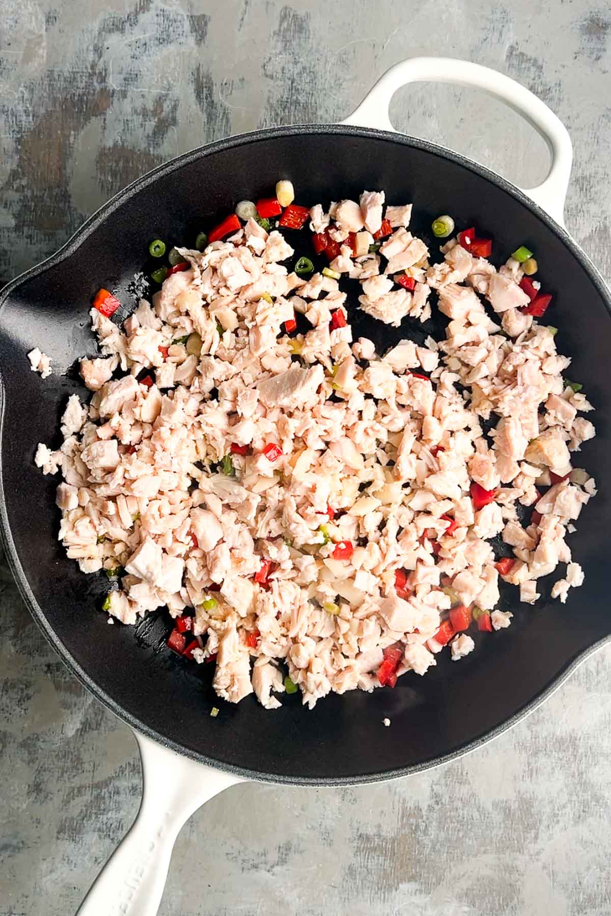 Chopped cooked chicken, diced red bell peppers, and green onions are being saut&eacute;ed in a black skillet with a white handle.