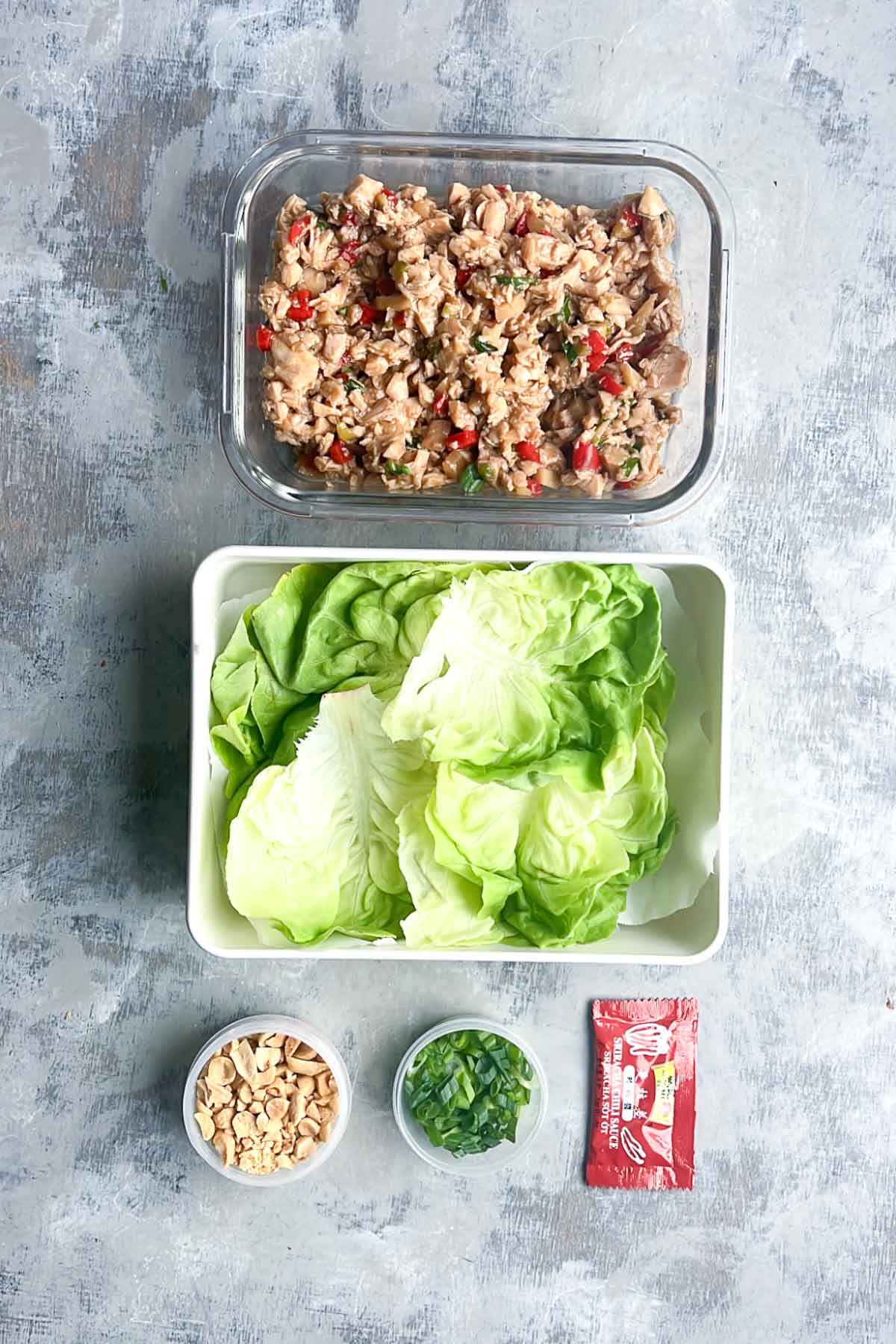 Overhead view of a meal kit for chicken lettuce cups, featuring cooked chicken and vegetables in a glass container, fresh lettuce leaves, chopped peanuts, sliced green onions, and a Sriracha packet arranged on a gray surface.