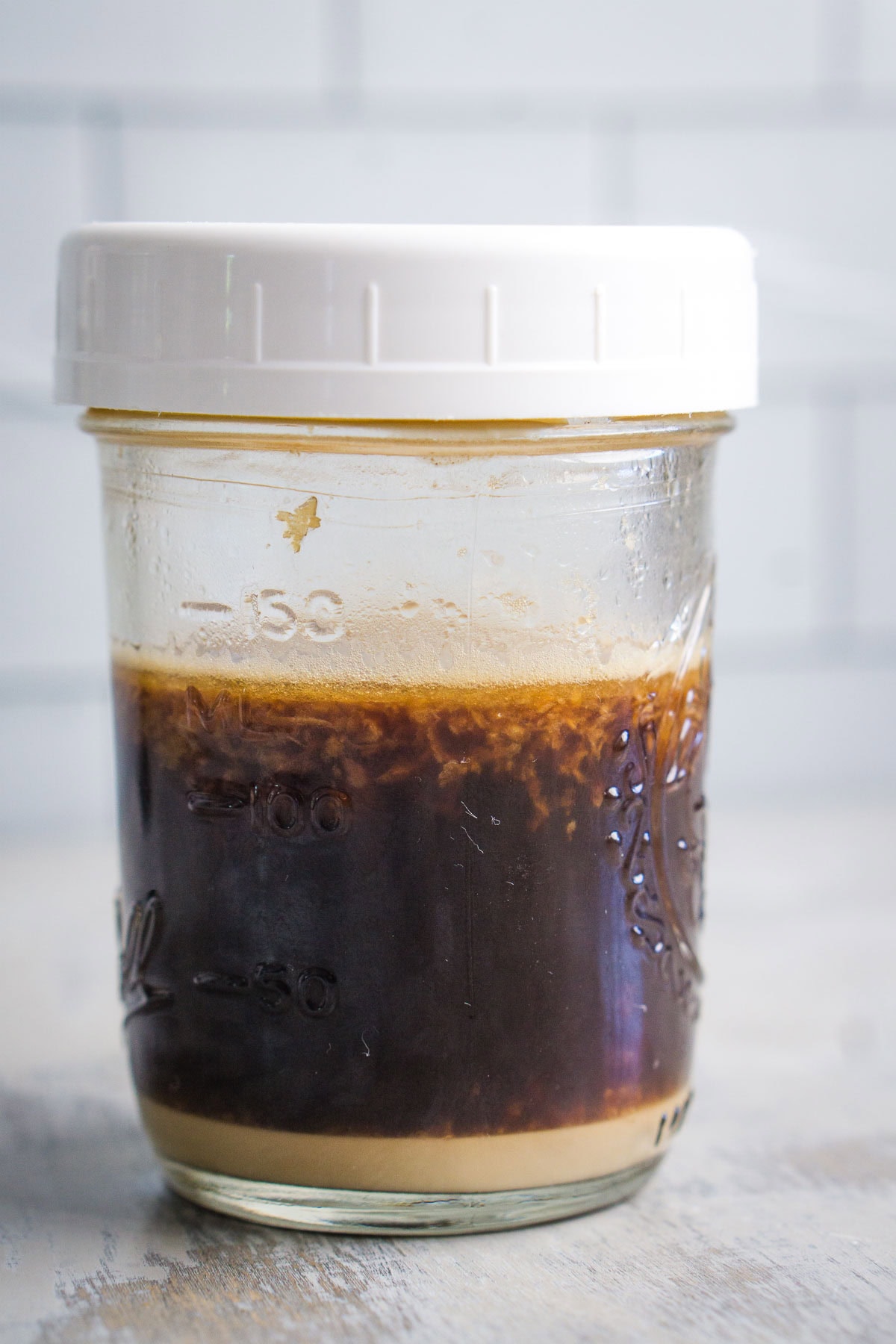 A glass jar with a white lid contains a dark brown liquid with a thick beige layer settled at the bottom. The jar is placed on a light surface against a tiled background.