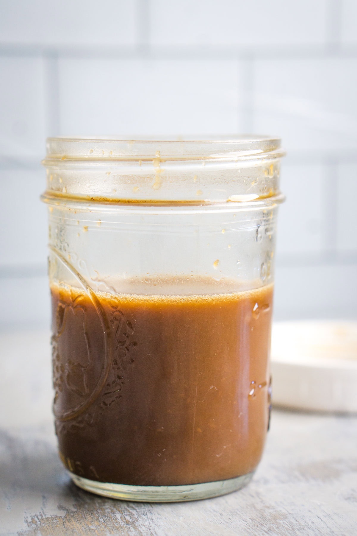 A glass mason jar filled with brown sauce sits on a light-colored surface with a white lid in the background.