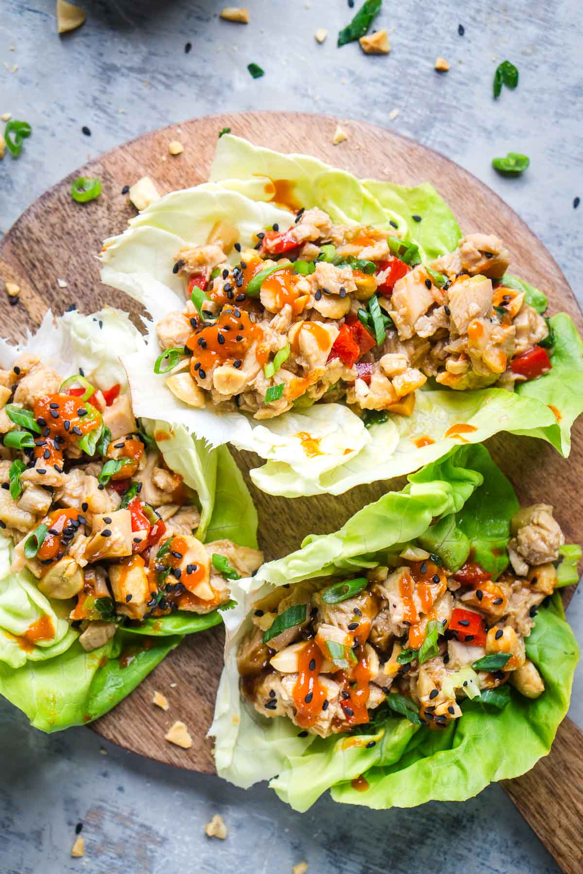 Three chicken lettuce wraps filled with chopped vegetables, diced chicken, and topped with crushed peanuts and chili sauce, served on a round wooden board.