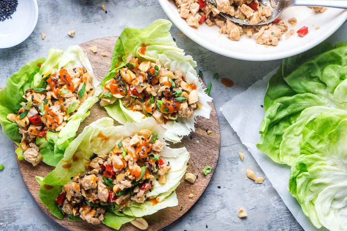 Three chicken lettuce wraps filled with diced vegetables and chicken, garnished with sauce and chopped nuts, are displayed on a round wooden board next to a bowl of filling and fresh butter lettuce leaves.