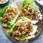 Three chicken lettuce wraps filled with chopped vegetables, diced chicken, and topped with crushed peanuts and chili sauce, served on a round wooden board.
