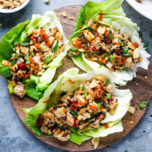 Three chicken lettuce wraps filled with chopped vegetables, diced chicken, and topped with crushed peanuts and chili sauce, served on a round wooden board.