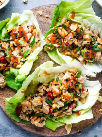 Three chicken lettuce wraps filled with chopped vegetables, diced chicken, and topped with crushed peanuts and chili sauce, served on a round wooden board.