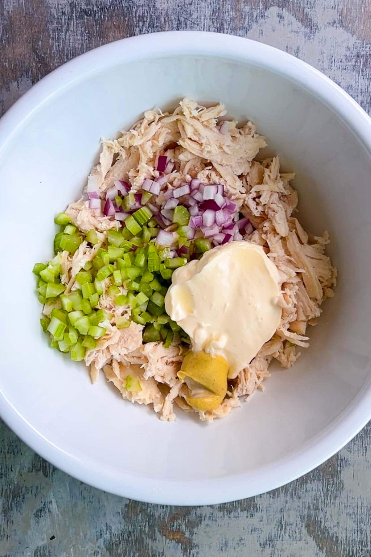 A white bowl containing chicken salad ingredients including shredded chicken, chopped celery, diced red onion, mayonnaise, and mustard on a gray surface.