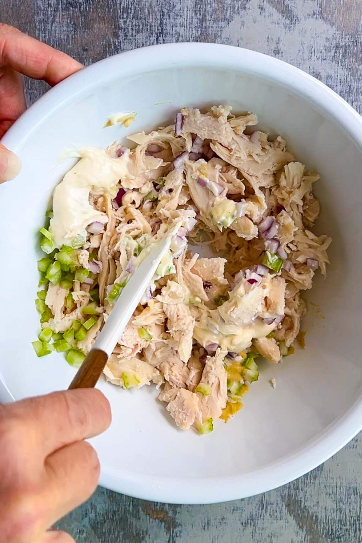 A hand mixing chicken salad with shredded chicken, chopped celery, red onion, and mayonnaise in a white bowl using a spatula.