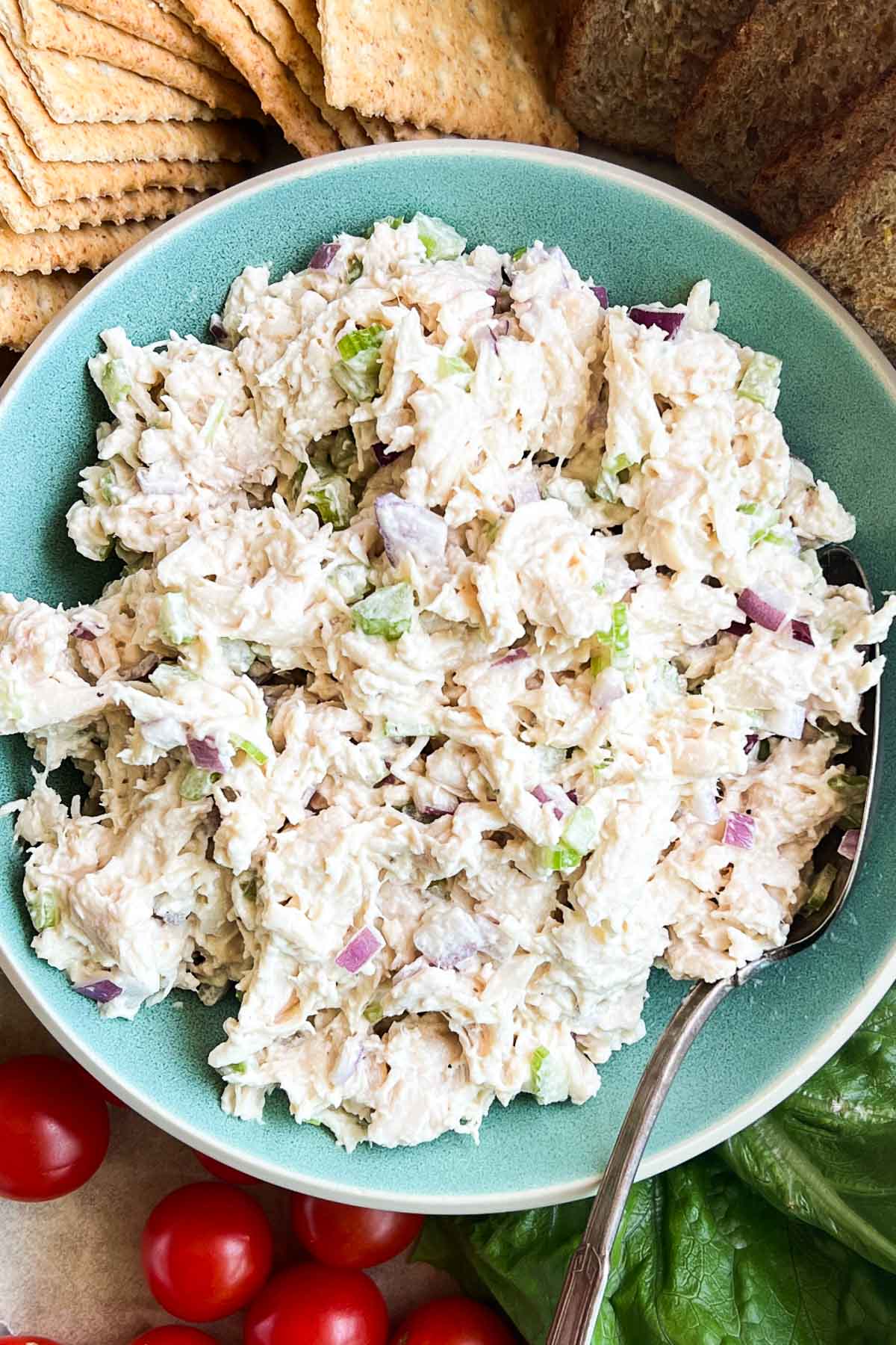 A bowl of chicken salad with diced celery and red onions is surrounded by crackers, bread slices, cherry tomatoes, and lettuce.