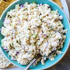 A bowl of classic chicken salad with chopped celery and onions is served alongside assorted crackers on a white plate.