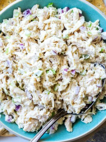 A bowl of classic chicken salad with chopped celery and onions is served alongside assorted crackers on a white plate.
