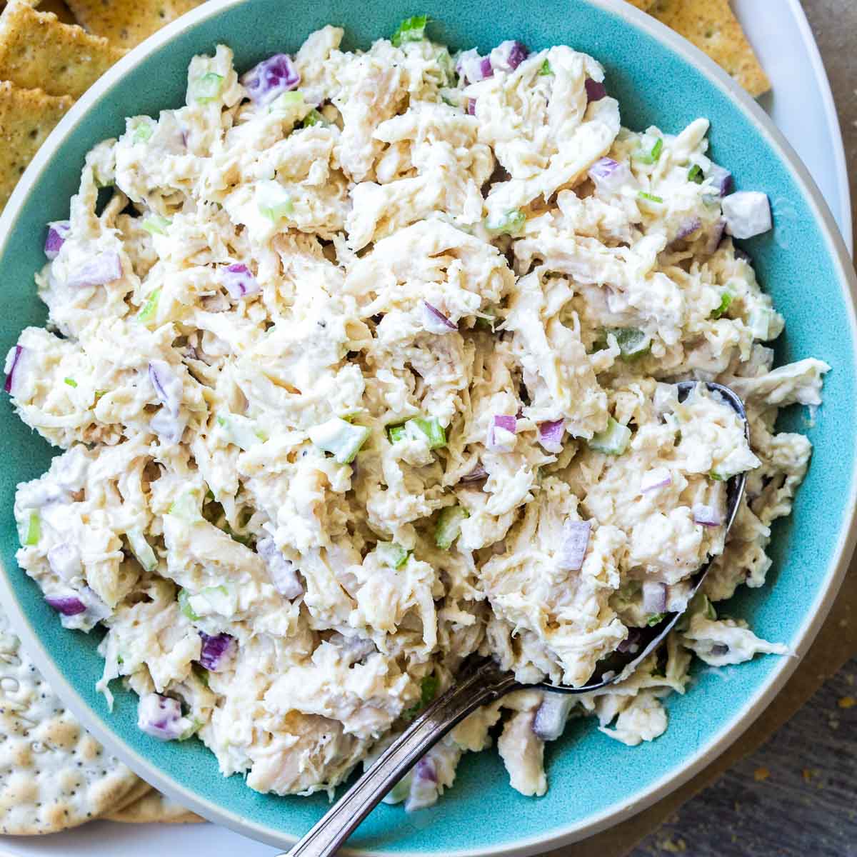 A bowl of classic chicken salad with chopped celery and onions is served alongside assorted crackers on a white plate.