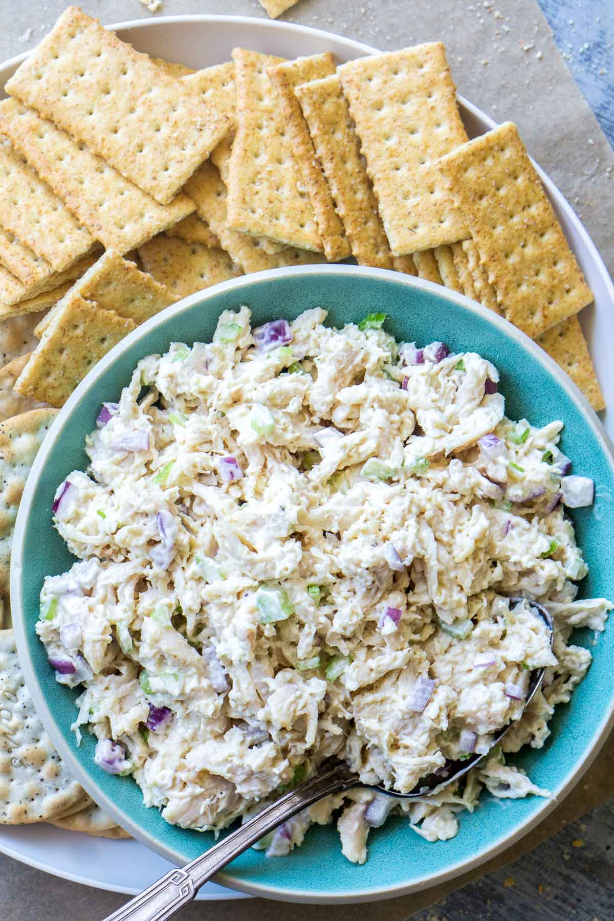 A bowl of classic chicken salad with chopped celery and onions is served alongside assorted crackers on a white plate.